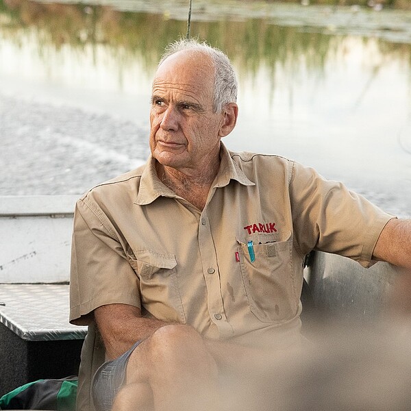 Älterer Mann im TARUK-Hemd sitzt auf einem Boot; im Hintergrund sind Wasser und ein bewaldetes Ufer zu sehen.