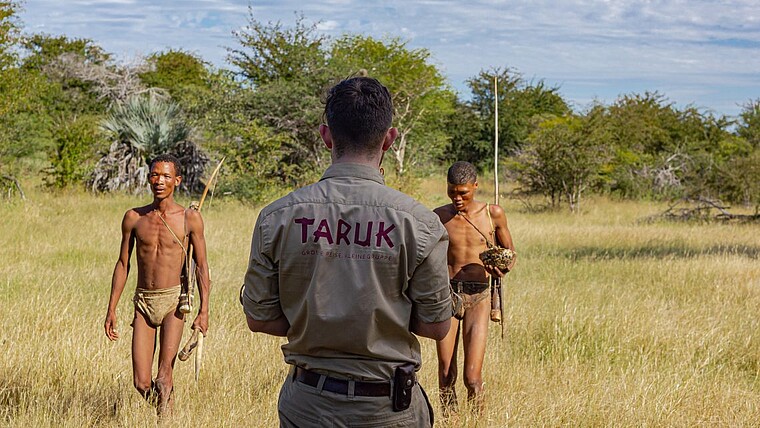 Eine Person mit TARUK-Hemd steht zwei traditionell gekleideten Menschen in einer trockenen Graslandschaft gegenüber.