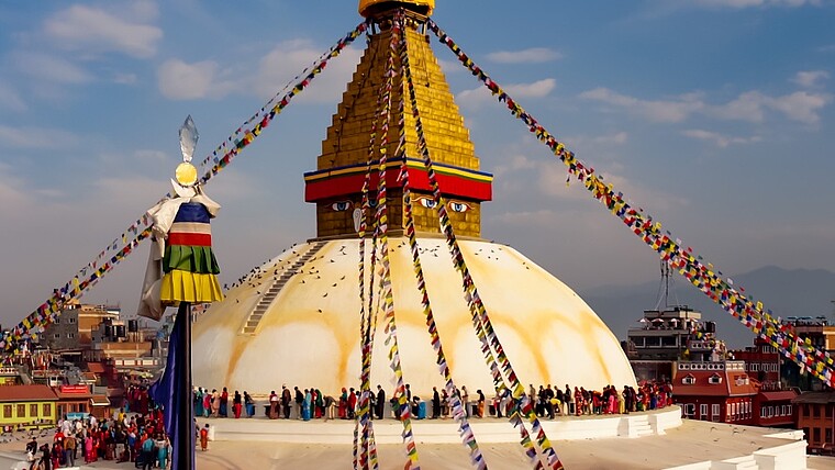Weiße Stupa mit goldener Spitze in Kathmandu umgeben von bunten Gebetsflaggen. Menschen stehen auf dem umlaufenden Balkon und schauen über die Stadt.