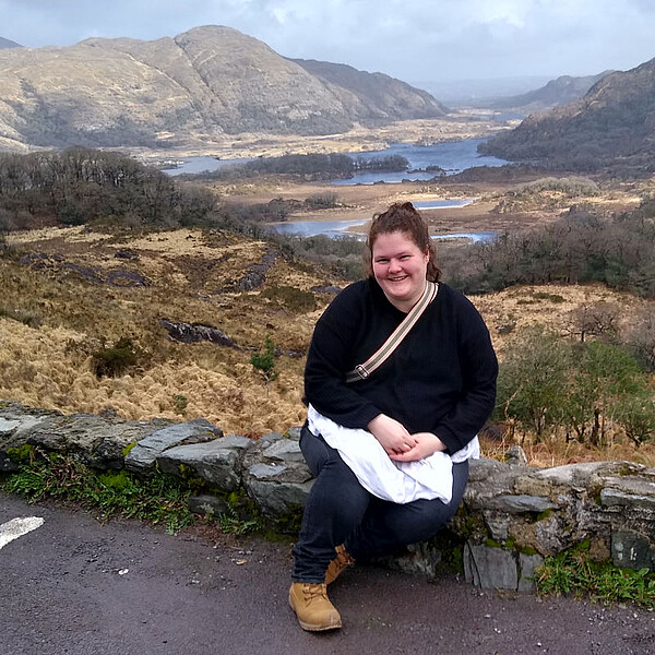 Vanessa Wendt sitzt lächelnd auf einer Steinmauer mit Blick auf hügelige Landschaft und Seen im Killarney Nationalpark in Irland.