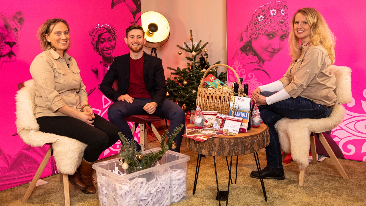 Drei Personen sitzen auf Stühlen. Ein kleiner Weihnachtsbaum steht neben einem runden Holztisch mit Geschenken, einem Korb und Werbematerialien. Im Hintergrund sind pinke Banner mit Gesichtern zu sehen.