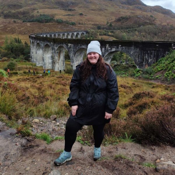 Vanessa Wendt sitzt lächelnd auf einer Steinmauer mit Blick auf hügelige Landschaft und Seen im Killarney Nationalpark in Irland.